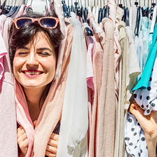 Eine Frau steckt den Kopf durch einen Kleiderständer und lacht in die Kamera.