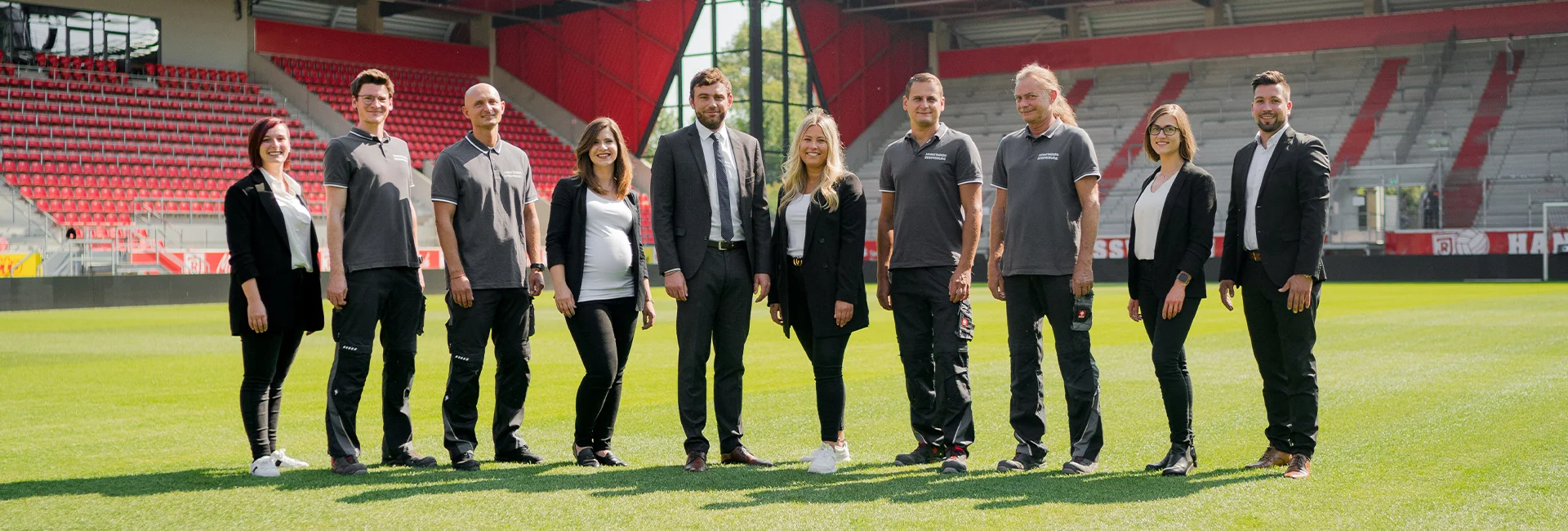 Das Team des Jahnstadions steht in einer Reihe auf dem Fußballfeld eines Stadions mit rot‑weißen Sitzreihen; einige tragen Anzüge, andere graue Poloshirts.