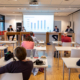 Man sieht vier Personen, die an braunen hohen Tischen auf eine Podium vor einer Leinwand mit einem Balkendiagramm sitzen sowie Menschen von hinten fotografiert, die ihnen zuschauen.