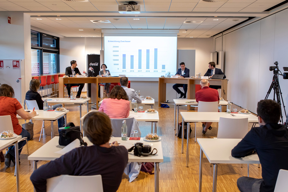 Man sieht vier Personen, die an braunen hohen Tischen auf eine Podium vor einer Leinwand mit einem Balkendiagramm sitzen sowie Menschen von hinten fotografiert, die ihnen zuschauen.