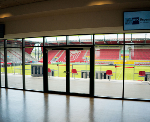 Besondere Ausblicke. Man sieht den Blick in den Innenbereich mit Rasen und Tribüne des Jahnstadions durch eine große bodentiefe Fensterfront.