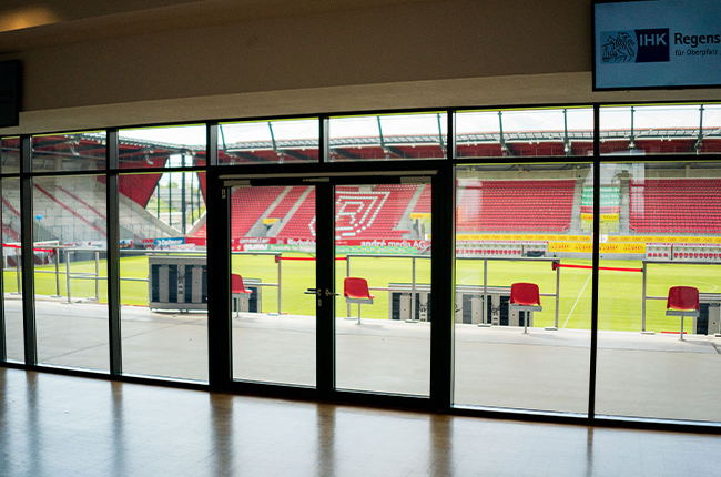 Besondere Ausblicke. Man sieht den Blick in den Innenbereich mit Rasen und Tribüne des Jahnstadions durch eine große bodentiefe Fensterfront.