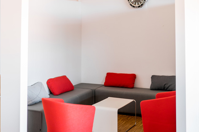 Man sieht eine schwarze Couch ohne Lehne mit roten und schwarzen Kisen und zwei rote Sessel sowie ein weißer Hocker als Tisch in der Mitte.
