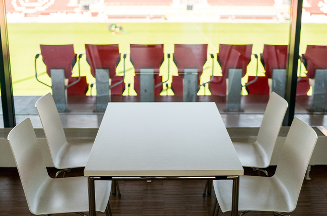 Man sieht einen weißen Tisch mit vier weißen Stühlen. Im Hintergrund schaut man durch ein Fenster ins Jahnstadion auf Rasen und Tribüne.