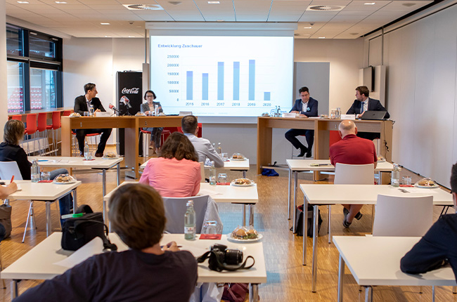 Man sieht vier Personen, die an braunen hohen Tischen auf eine Podium vor einer Leinwand mit einem Balkendiagramm sitzen sowie Menschen von hinten fotografiert, die ihnen zuschauen.