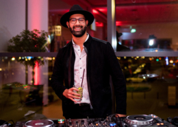 Man sieht einen lachenden Mann mit Hut, schwarzer Jacke und weißem Hemdn, der einen Cocktail in der Hand hält und an einem Mischpult steht.