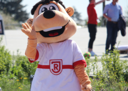 Man sieht das Maskottchen des SSV Jahn Regensburg, es trägt ein weißes T-Shirt mit roten Elementen und dem Vereinswappen. Es ist ein Mischung zwischen Mensch und Bär und kann nicht genau definiert werden. Es winkt in die Kamera.