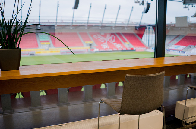 Man sieht einen Stehtisch vor einem Fenster durch das man auf den Innenraum des Jahnstadions sieht.