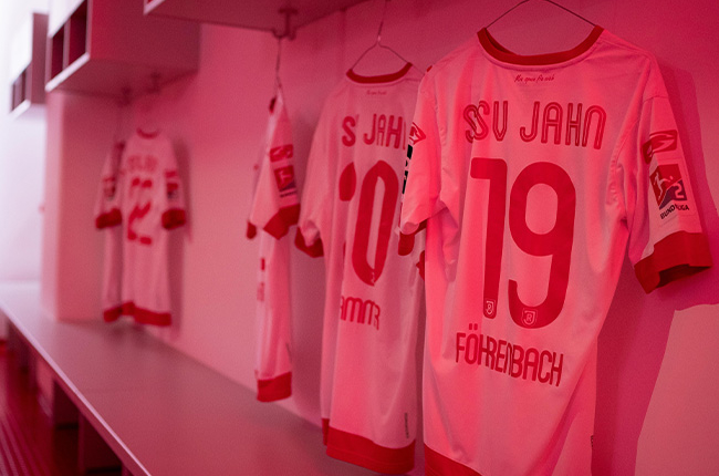 Man sieht verschiedene Trikots des SSV Jahn Regensburg in der Gaderobe hängen, das Foto ist in rötlichem Farbton aufgenommen.