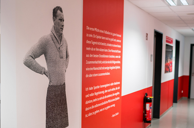 Man sieht einen Gang im Jahnstadion. An der Wand hängen Bilder und die Wand ist bis zur Hälfte rot gestrichen. Groß ist eine schwarz-weiß Fotografie eines Mannes abgebildet sowie ein Text daneben.