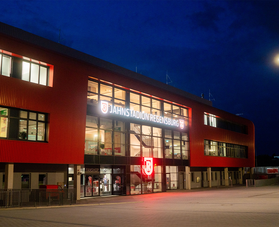 Man sieht ein Foto des Eingangsbereichs des Jahnstadion Regensburg bei Nacht mit leuchtendendem Schriftzug.