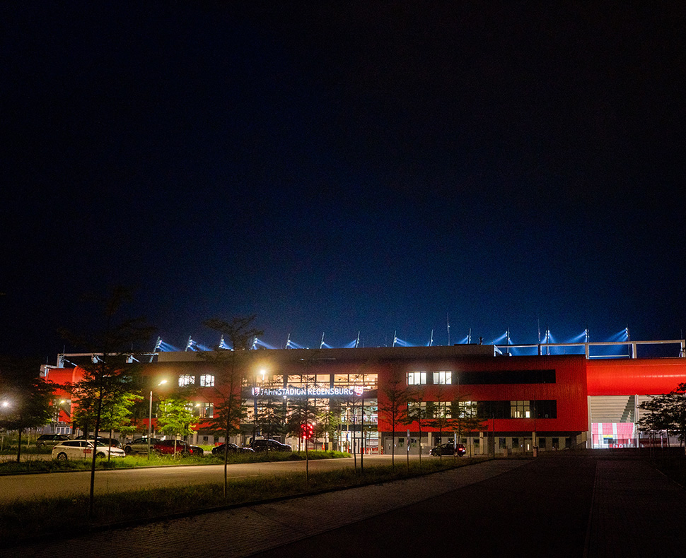 Man sieht ein Foto vom Jahnstadion Regensburg bei Nacht mit leuchtender Flutlichtanlage vom Parkplatz aus.