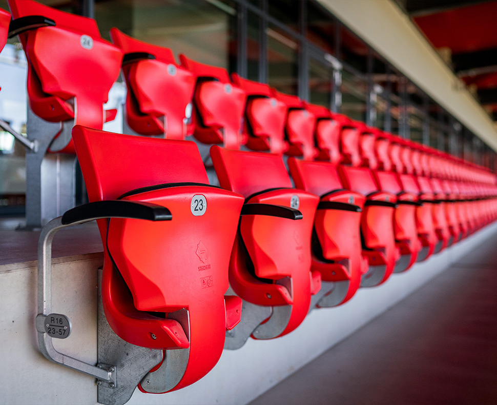Man sieht eine Nahaufnahme von roten Klappstühlen in der Tribüne eines Fußballstadions.