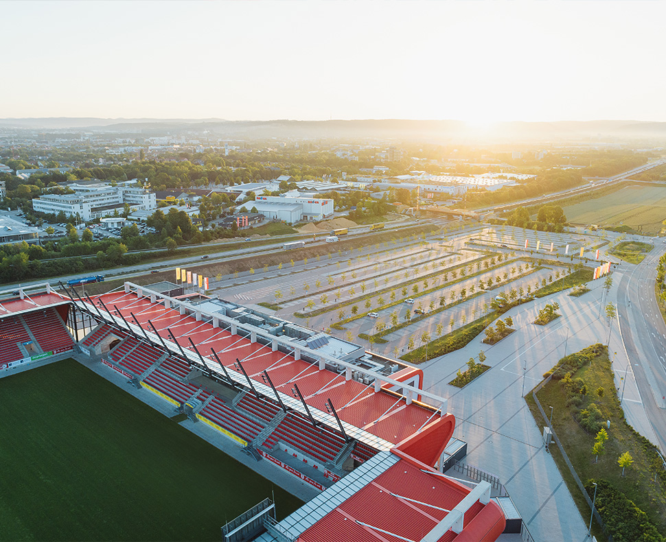 Man sieht eine Luftaufnahme eines großen Parkplatzes sowie nebenan das Jahnstadion bei Sonnenaufgang.