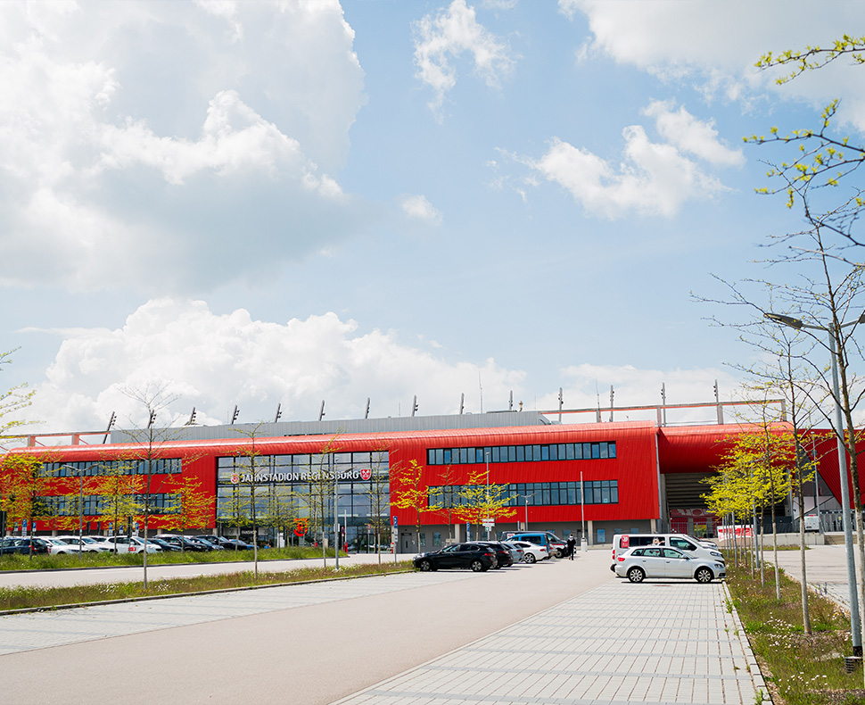 Man sieht einen Parkplatz mit wenigen Autos und im Hintergrund das Jahnstadion Regensburg bei Tag.