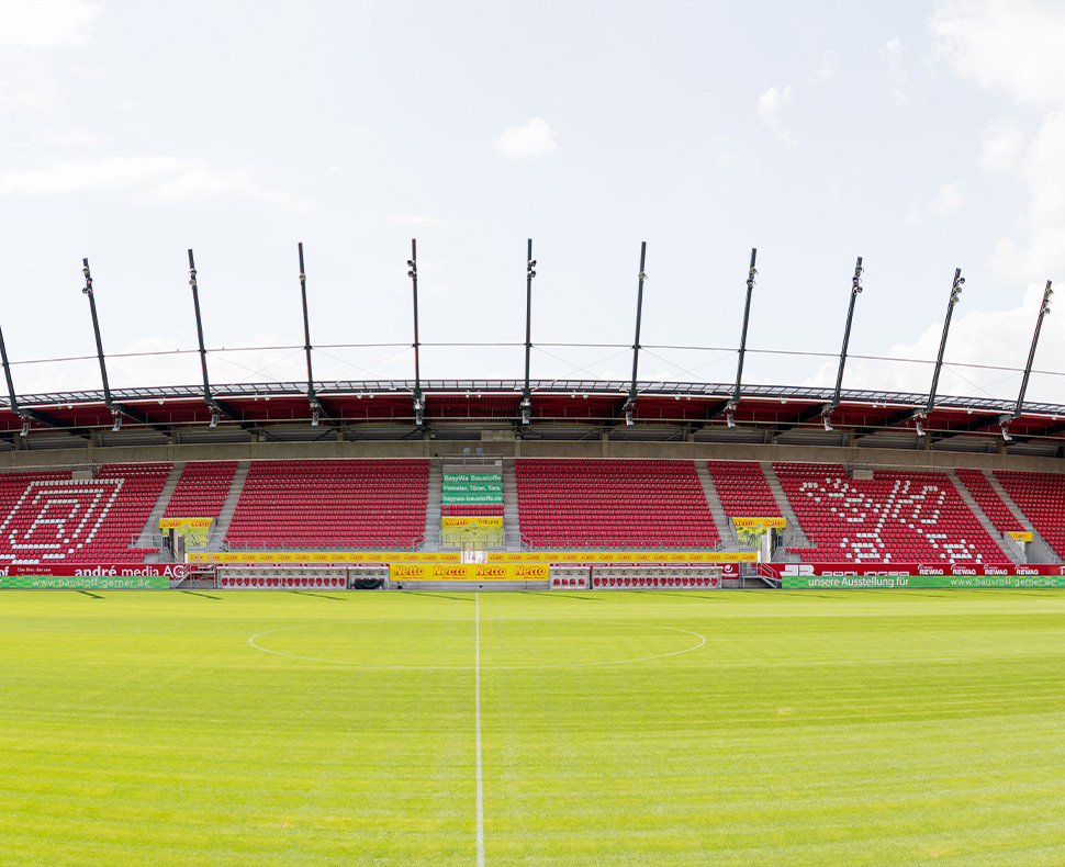 Man sieht ein Foto der Rasenfläche des Jahnstadions sowie einer Tribüne auf der Längsseite mit roten Sitzen und dem Wappen der Stadt Regensburg sowie des Jahnregensburg in weiß.