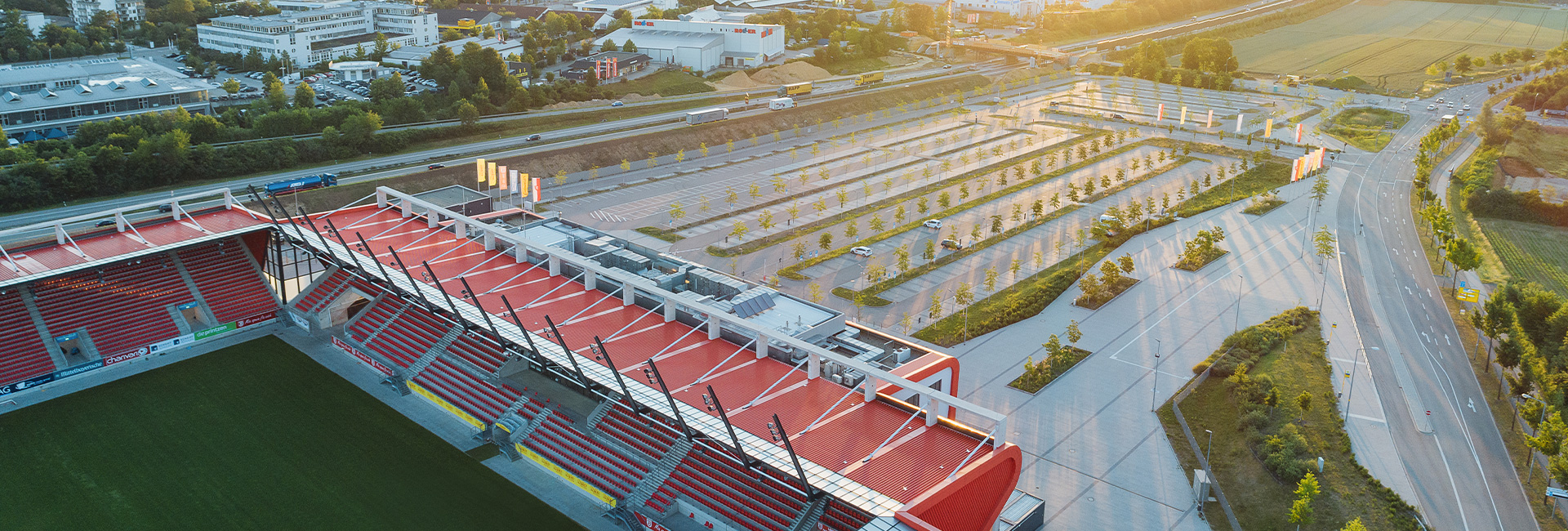 Man sieht eine Luftaufnahme eines großen Parkplatzes sowie nebenan das Jahnstadion bei Sonnenaufgang.