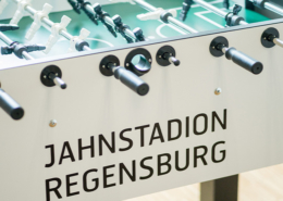 Man sieht einen weißen Kickerkasten in der Nahaufnham auf dem Jahnstadion Regensburg in schwarz geschrieben ist.