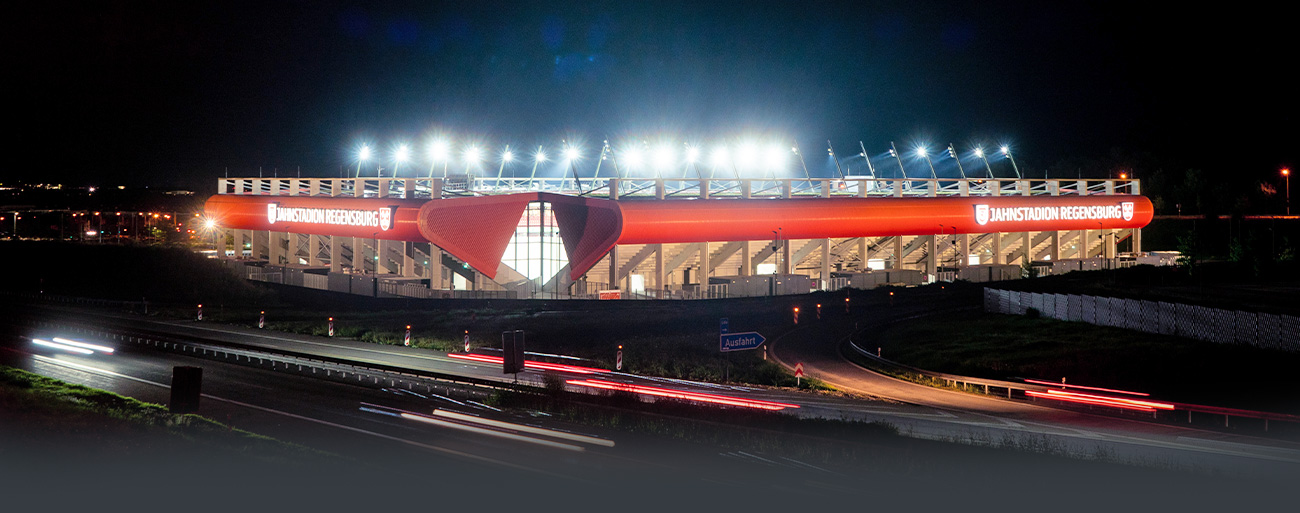 Man sieht ein Foto vom Jahnstadion Regensburg bei Nacht mit leuchtender Flutlichtanlage von weiter weg.