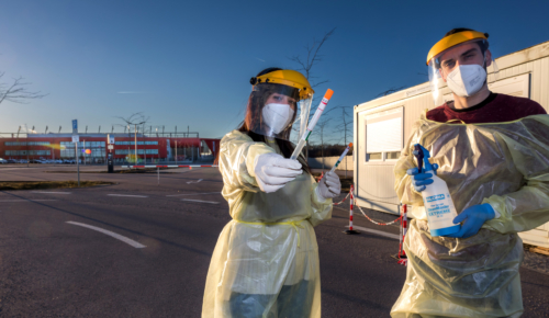 Man sieht zwei Personen in Schutzanzügen und Maske die Coronatestungen auf dem Parkplatz des Jahnstadions durchführen.