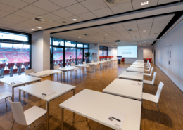 Man sieht einen große Raum mit Blick auf den Innenbereich des Jahnstadions, in dem viele weiße Tische mit jeweils einem weißen Stuhl in U-Form aufgebaut sind.