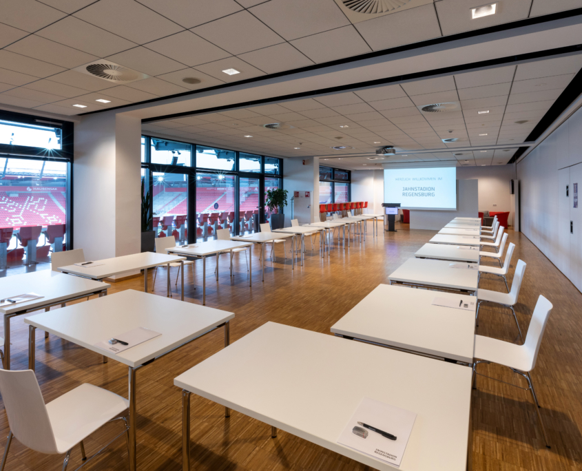 Man sieht einen große Raum mit Blick auf den Innenbereich des Jahnstadions, in dem viele weiße Tische mit jeweils einem weißen Stuhl in U-Form aufgebaut sind.
