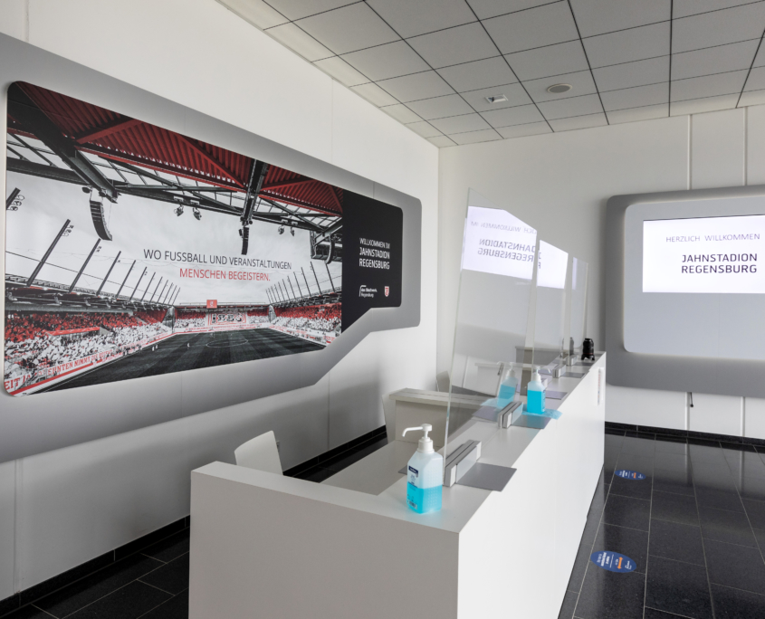 Man sieht den weißen Eingangstresen im Jahnstadion auf dem Desinfektionsmittelspender stehen und eine Plexiglasscheibe aufgebaut ist.