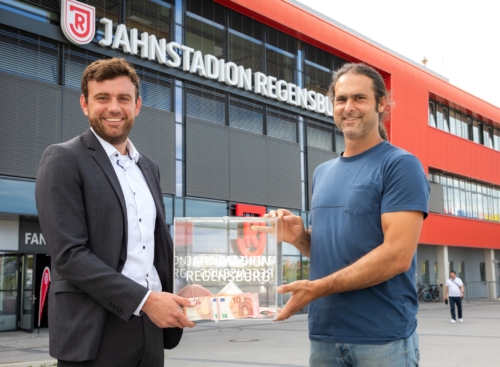 Man sieht zwei Männer die sich vor dem Jahnstadion einen durchsichtige Box mit Geldscheinen übergeben.