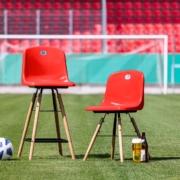 Man sieht zwei unterschiedlich hohe rohe Stühle vor einem Fußballtor und einer Tribüne mit roten Sitzen. Daneben liegen ein Fußball und ein Glas mit Bier.