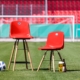 Man sieht zwei unterschiedlich hohe rohe Stühle vor einem Fußballtor und einer Tribüne mit roten Sitzen. Daneben liegen ein Fußball und ein Glas mit Bier.