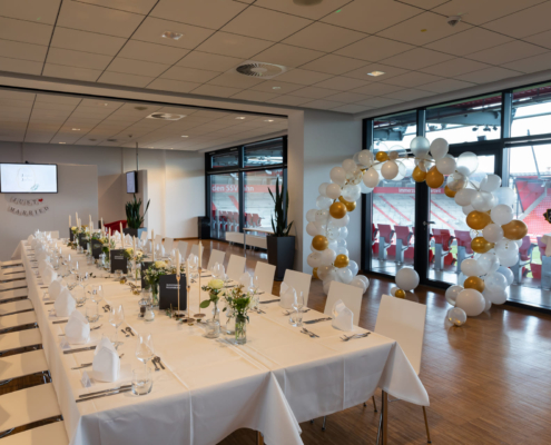 Man sieht einen Raum im Jahnstadion mit einer festlich gedeckten Tafel mit weißer Tischdecke und viel Dekoration. Im Hintergrund ist ein Dekobogen aus weißen und goldenen Luftballons zu sehen. Durch eine Fensterfront kann man ins Jahnstadion sehen.