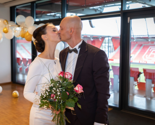 Man sieht einen Mann im schwarzen Anzug und eine Frau in einem weißen Kleid mit einem Blumenstrauß. Die beiden küssen sich. Im Hintergrund blickt man durch ein großes bodentiefes Fenster auf die Tribüne des Jahnstadions.
