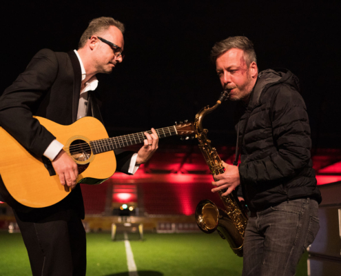 Man sieht zwei Männer, die Posaune und Gitarre spielen. Im Hintergrund sieht man Umrisse eines Fußballstadions in der Nacht.