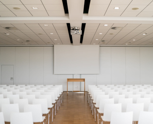 Man sieht einen Konferenzraum in dem links und rechts weiße Stuhlreihen aufgebaut sind. An der Wand hängt eine weiße Leinwand.