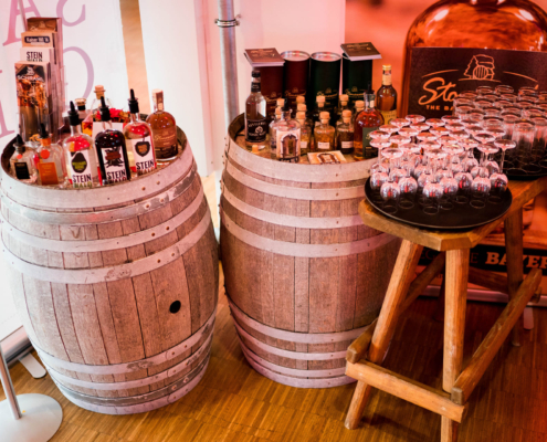 Man sieht zwei Holzfässer und einen Holztisch auf denen verschiedene Whiskeyflaschen und Gläser aufgebaut sind.