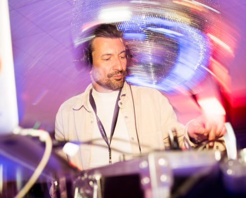Man sieht einen Mann an einem DJ Pult mit einer verschwommen Diskokugel im Hintergrund.