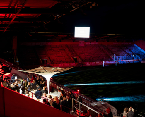 Man sieht eine Tribüne des Jahnstadions in der Nacht im Hintergrund sowie eine Gruppe Menschen auf einer Terrasse ins Stadion im Vordergrund.