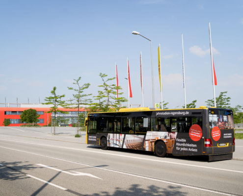 Man sieht einen Linienbus, der mit Werbung des Jahnstadions beklebt ist und am Jahnstadion vorbeifährt.