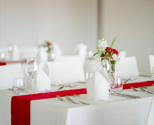Man sieht einen Tisch mit weißer Tischdecke, Besteck, Servietten, Gläsern, einem roten Tischläufer in der Mitte und rot-weißer Blumendeko.