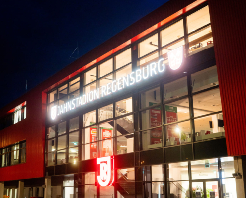 Man sieht ein Foto des Eingangsbereichs des Jahnstadion Regensburg bei Nacht mit leuchtendendem Schriftzug.