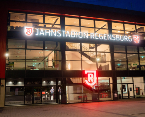 Man sieht ein Foto des Eingangsbereichs des Jahnstadion Regensburg bei Nacht mit leuchtendendem Schriftzug.