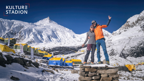 Man sieht einen Mann und eine Frau, die sich im Arm halten. Im Hintergrund sind gelb-blaue Zelte und schneebedeckte hohe Berge und ein Gipfel.