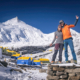 Man sieht einen Mann und eine Frau, die sich im Arm halten. Im Hintergrund sind gelb-blaue Zelte und schneebedeckte hohe Berge und ein Gipfel.