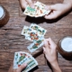 Man sieht zwei Hände, die Spielkarten in der Hand haben und Schafkopf spielen sowie einen Maßkrug mit Bier auf einem Holztisch.