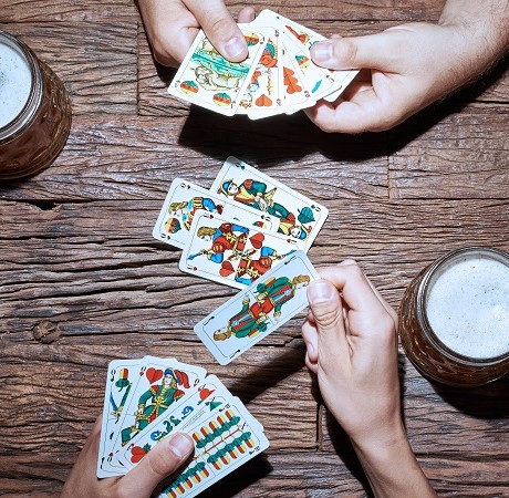 Man sieht zwei Hände, die Spielkarten in der Hand haben und Schafkopf spielen sowie einen Maßkrug mit Bier auf einem Holztisch.