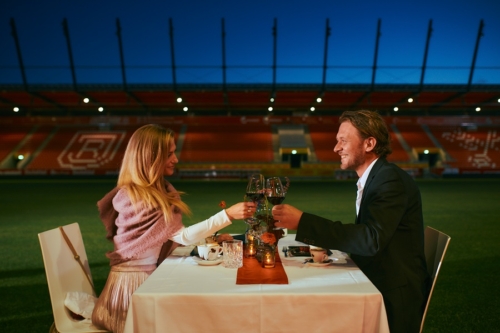 Man sieht eine Frau und einen Mann, beide elegant gekleidet, die an einem gedeckten Tisch im Jahnstation bei Nacht mit einem Glas Rotwein anstoßen.