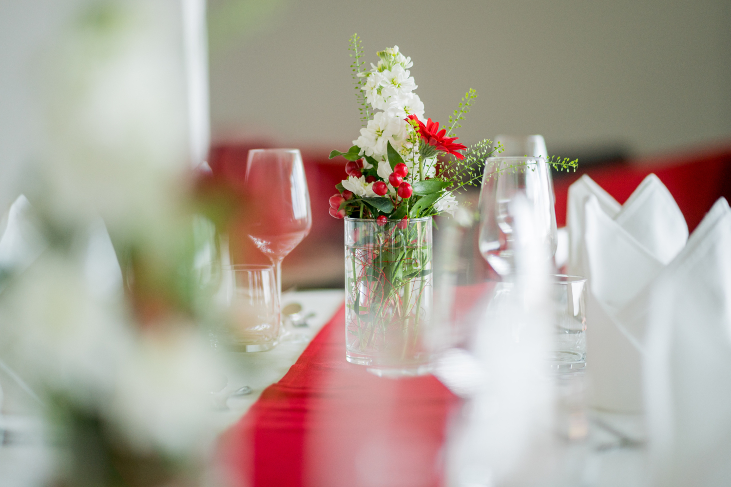 Man sieht ein Foto von einem gedeckten Tisch mit weißer und roter Tischdecke, Gläsern und einem Blumenbouquet in der Mitte.
