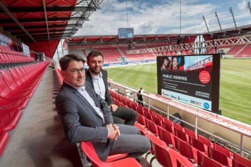 Man sieht ein Foto von zwei Männer die aufder Tribüne sitzen mit einem einer Leinwand zum Public Viewing.