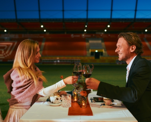 Man sieht eine Frau und einen Mann, die an einem Tisch sitzen, der auf dem Spielfeld des Stadions steht, beide stoßen mit ihren Gläsern an.