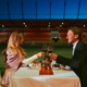 Man sieht eine Frau und einen Mann, die an einem Tisch sitzen, der auf dem Spielfeld des Stadions steht, beide stoßen mit ihren Gläsern an.